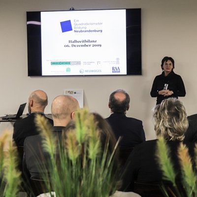 Eine Frau steht vor einer Gruppe neben ihr wird eine Präsentation abgespielt